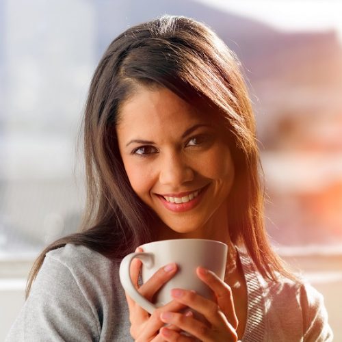 Woman drinking coffee at home with sunrise streaming in through window and creating flare into the lens.